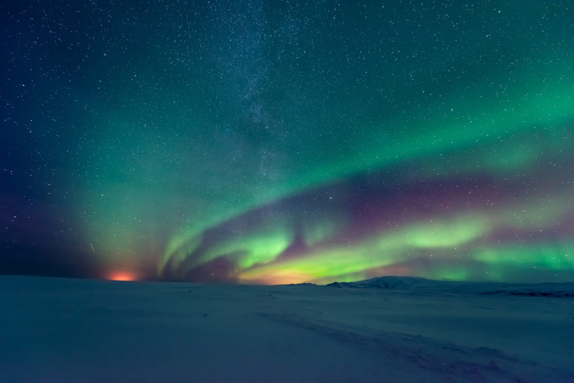 Vibrant green and purple aurora borealis swirl across a star-studded teal night sky, with a faint Milky Way band. A vast, snow-covered plain stretches below, meeting distant dark mountains and a warm orange glow on the left horizon.