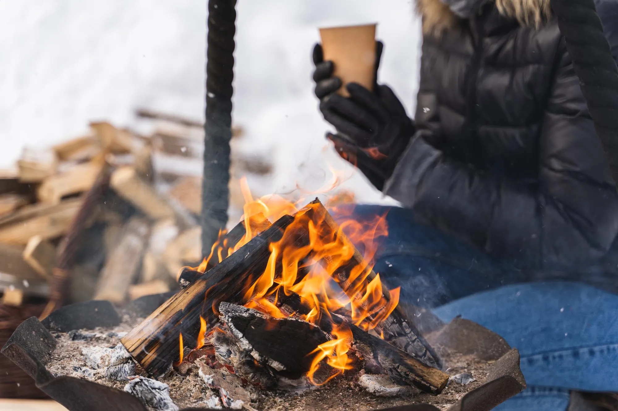 A person in a black puffer jacket and blue jeans sits by a metal fire pit, warming their gloved hands around a light brown paper cup. Bright orange and yellow flames consume dark logs and embers in the foreground. In the blurred background, a pile of stacked firewood is visible under a white, overcast sky.