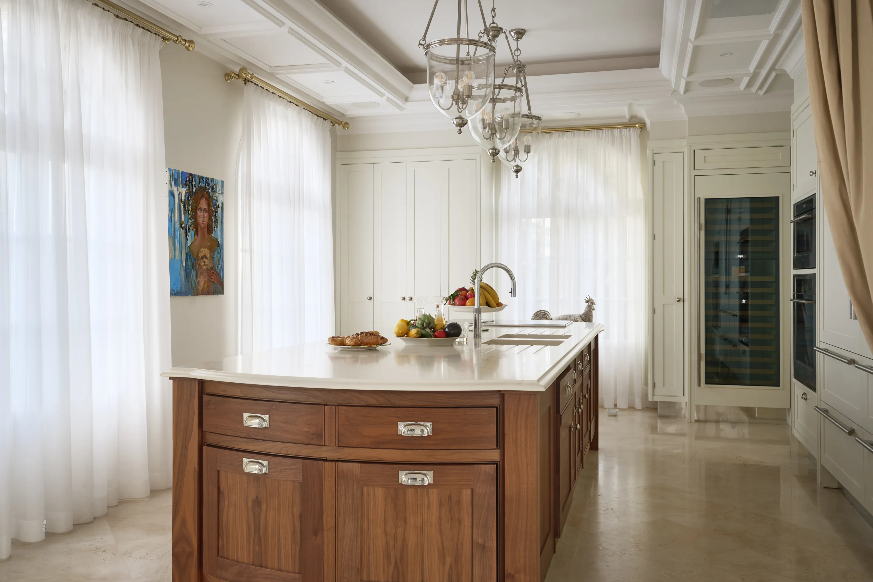 A bright, elegant kitchen features a central wooden island with a light countertop, a sink, fresh fruit, and pastries. White paneled cabinets line the walls, with a built-in wine cooler and ovens on the right. Large windows with sheer white curtains and gold rods are on the left, alongside a colorful painting. Three glass and silver pendant lights hang from the white coffered ceiling, illuminating the polished light marble floor.