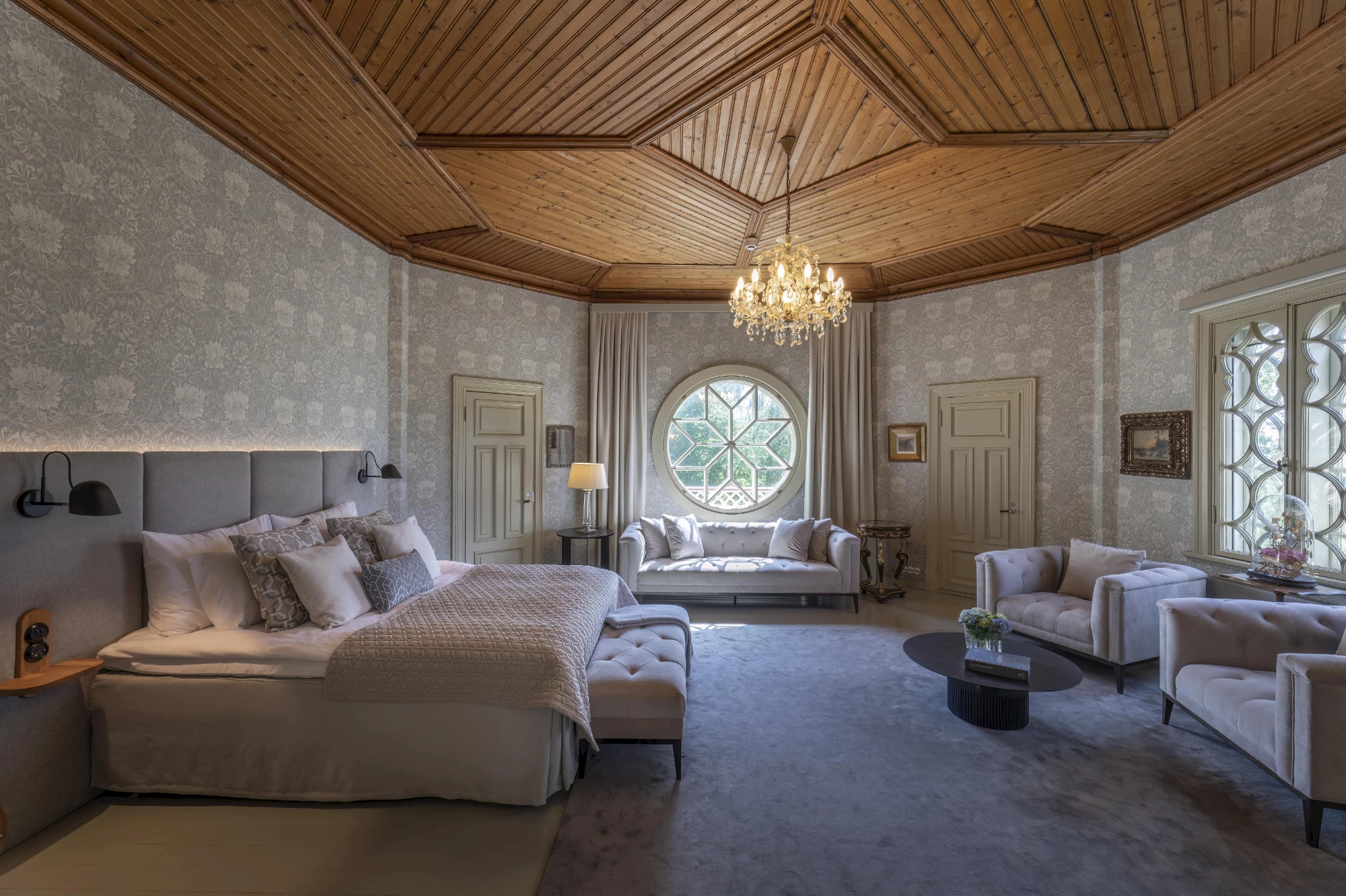 A spacious, elegant room features a striking wooden paneled ceiling with a complex geometric design, from which a crystal chandelier hangs. The walls are covered in light gray wallpaper with a subtle floral pattern. A large bed with a gray upholstered headboard, backlit by a strip light, is positioned on the left, flanked by black wall-mounted lamps. The bed is dressed in white linens, light gray pillows, and a quilted throw, with a light gray upholstered bench at its foot. The room boasts two distinct windows: a large circular window with a star-shaped grid on the far wall, and a tall rectangular window with an intricate curvilinear grid on the right, both framed by light beige drapes. Two light beige paneled doors are visible. Seating areas include a light beige tufted sofa near the circular window and two matching armchairs arranged around a dark oval coffee table with flowers and books near the rectangular window. The floor is covered in a soft light gray carpet.