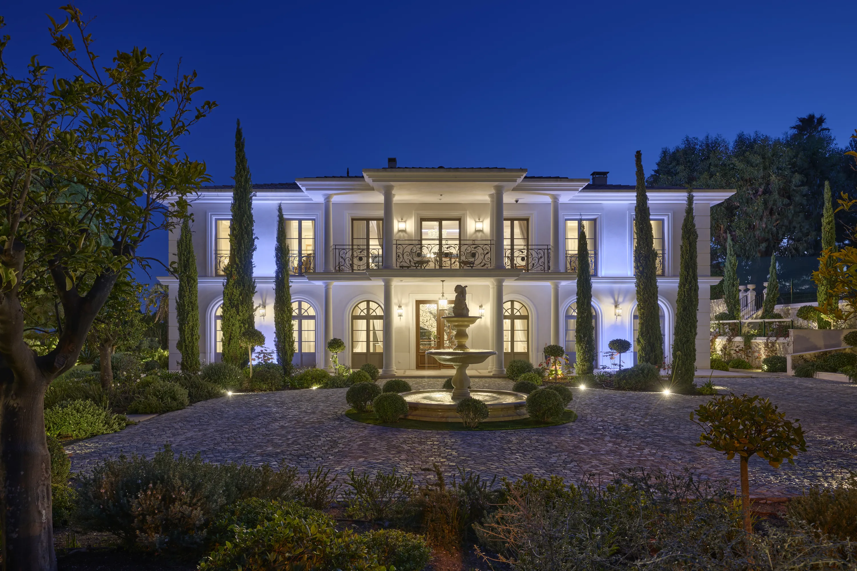 A stately white mansion with classical architecture stands brightly illuminated at twilight, featuring a central stone fountain in a circular cobblestone driveway. Tall, slender cypress trees and lush green foliage frame the property, all lit by a combination of warm interior lights, cool blue uplighting on the facade, and numerous ground spotlights. The deep blue sky above completes the serene evening scene.