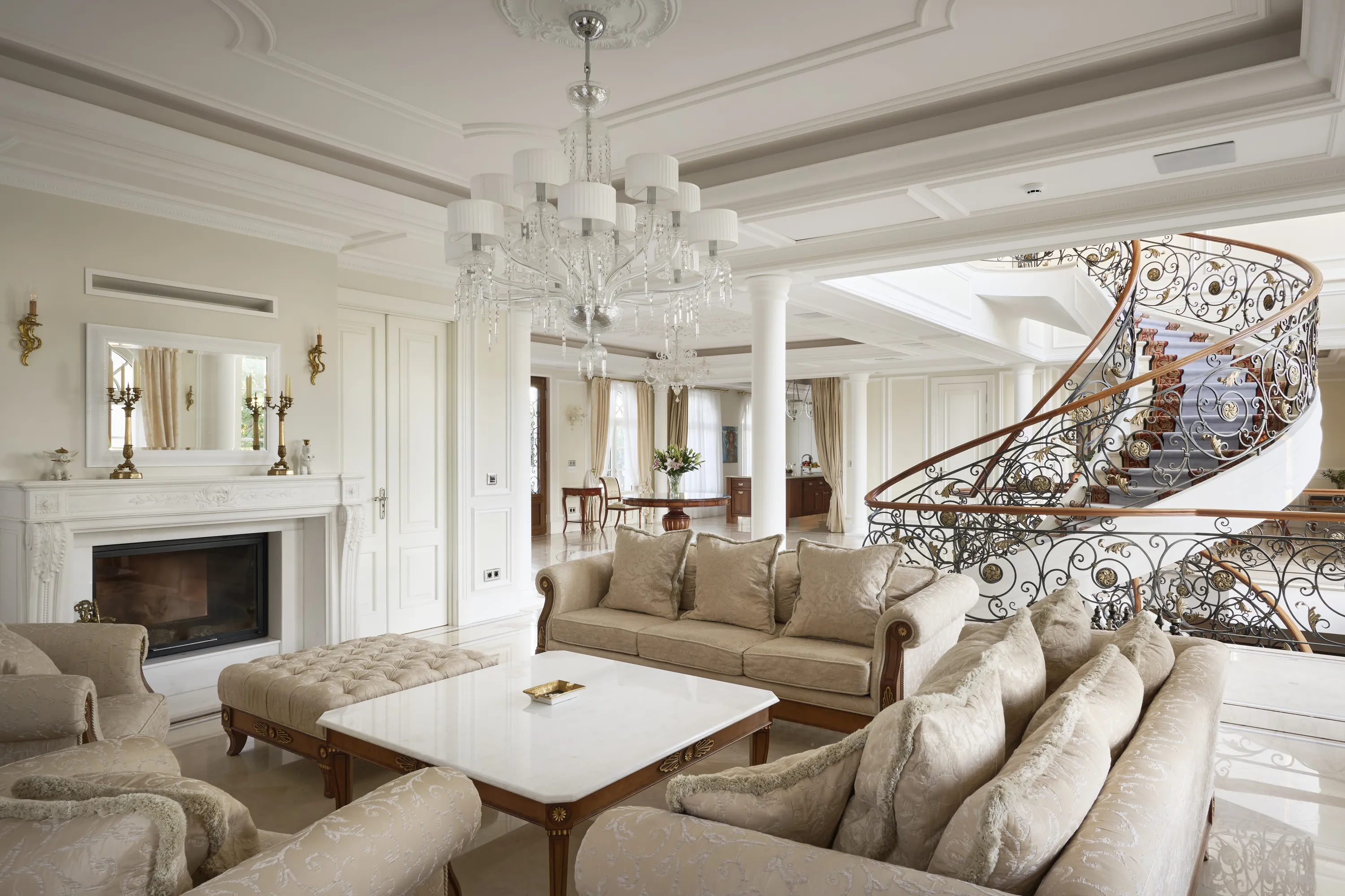 A spacious, opulent living room features light beige sofas and armchairs arranged around a white marble coffee table and a tufted ottoman. A grand white fireplace with a mirror above it is flanked by brass wall sconces. Overhead, an elaborate crystal chandelier with white shades illuminates the room, which boasts intricate white ceiling moldings. To the right, a magnificent spiral staircase with dark iron scrollwork and gold accents, a wooden handrail, and a patterned grey carpet ascends. The room has polished light floors and large windows with cream curtains, creating a bright and elegant atmosphere.