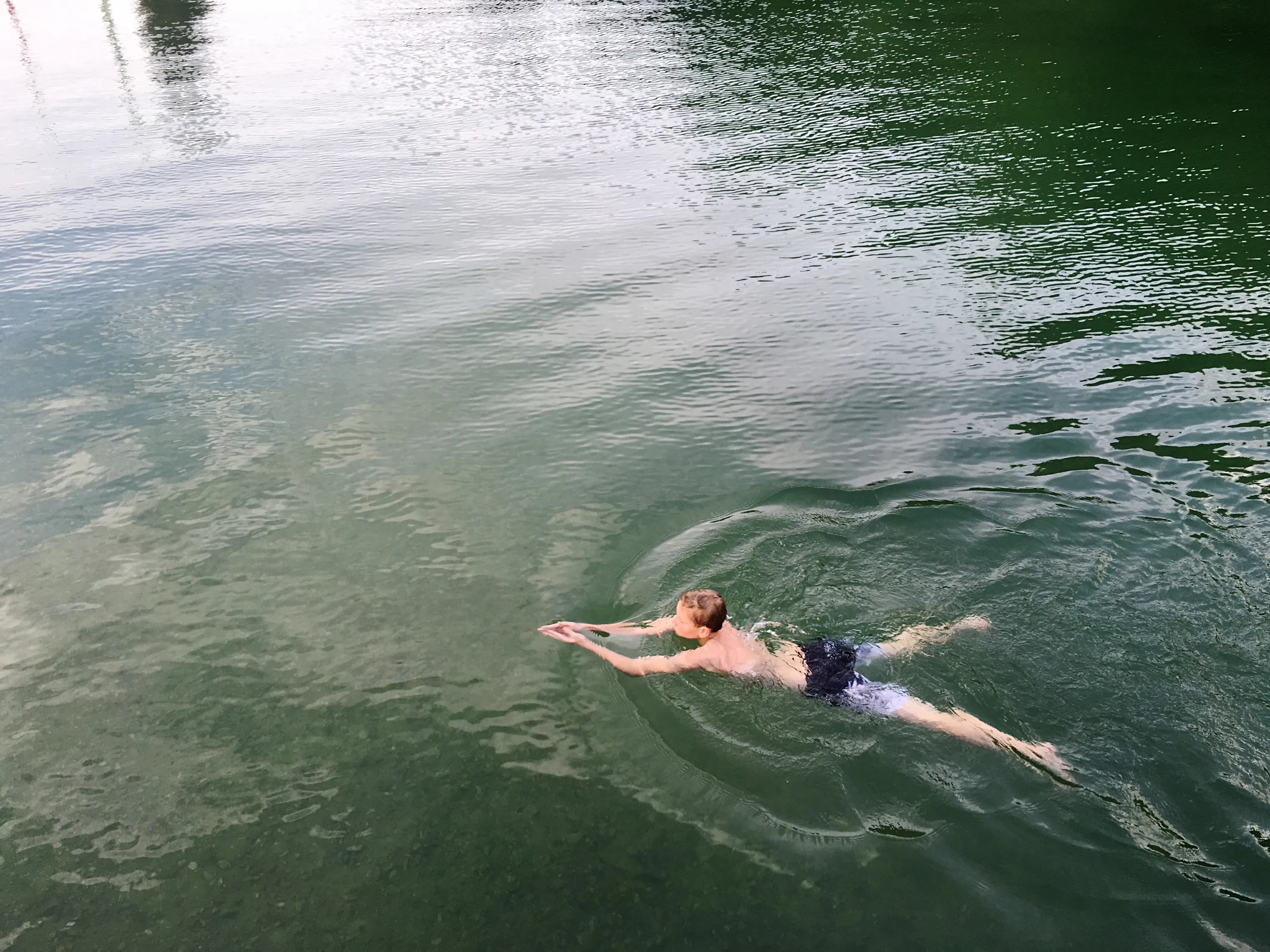 A person swims on their stomach in a body of water, with arms extended and legs kicking. The water displays ripples and reflections, transitioning from lighter greenish-blue on the left, where the bottom is visible, to a darker green on the right.