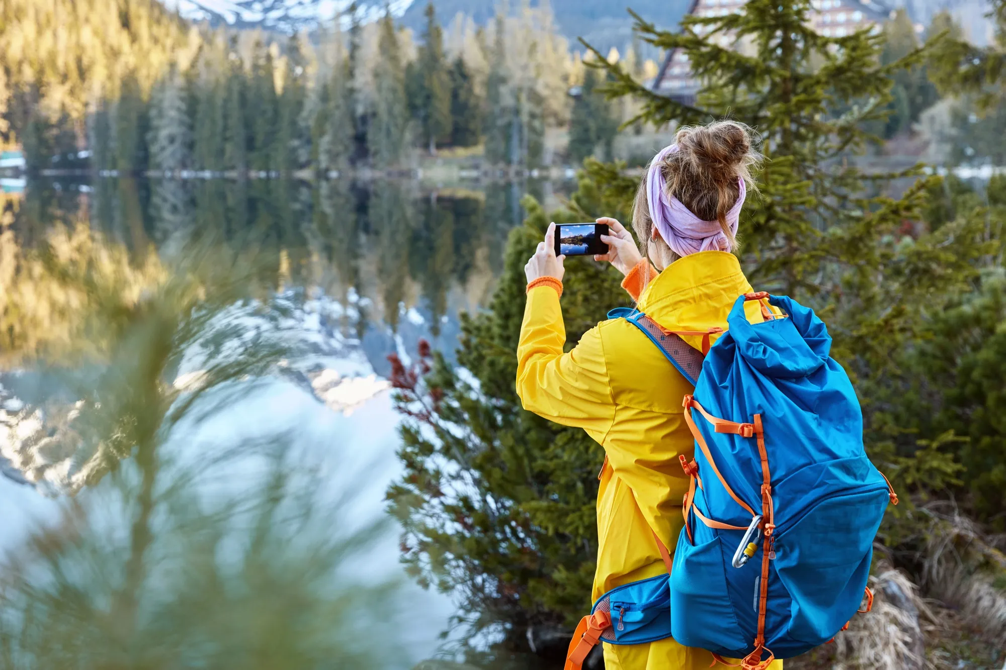 A person, viewed from behind, wears a bright yellow jacket and a blue backpack with orange straps, holding a smartphone horizontally to photograph a scenic lake. Their hair is tied in a bun with a lavender headband. The calm lake reflects a dense forest of evergreen trees and distant snow-capped mountains under bright, natural daylight.