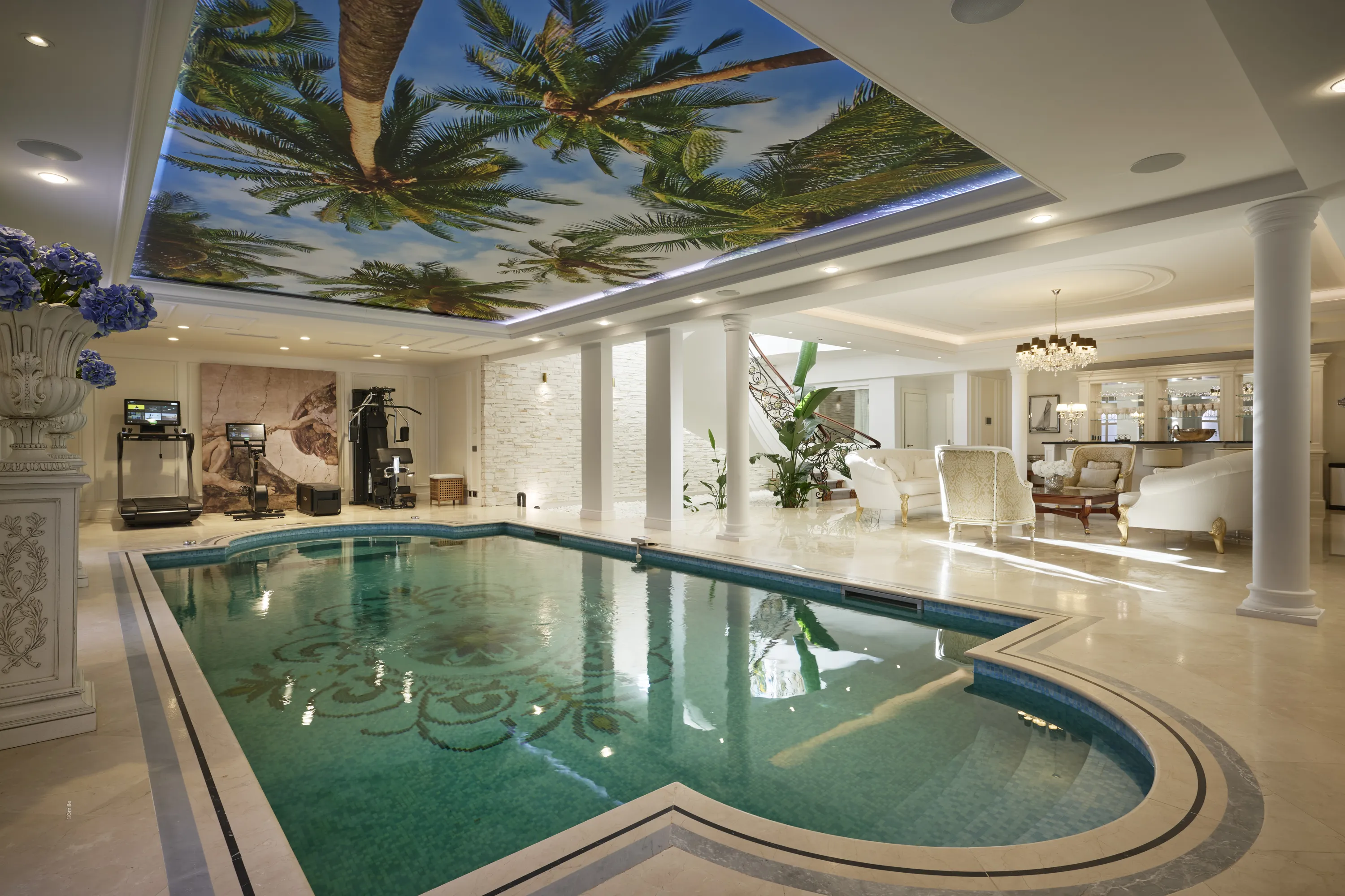A luxurious indoor swimming pool area features a large ceiling light panel displaying a vibrant palm tree and blue sky mural. The teal pool has mosaic patterns on its bottom, bordered by light marble with dark stripes. To the left, a gym area includes black exercise equipment and a large wall art print. To the right, a lounge area with white and cream upholstered furniture, a bar, and a crystal chandelier is illuminated by bright natural and artificial light.