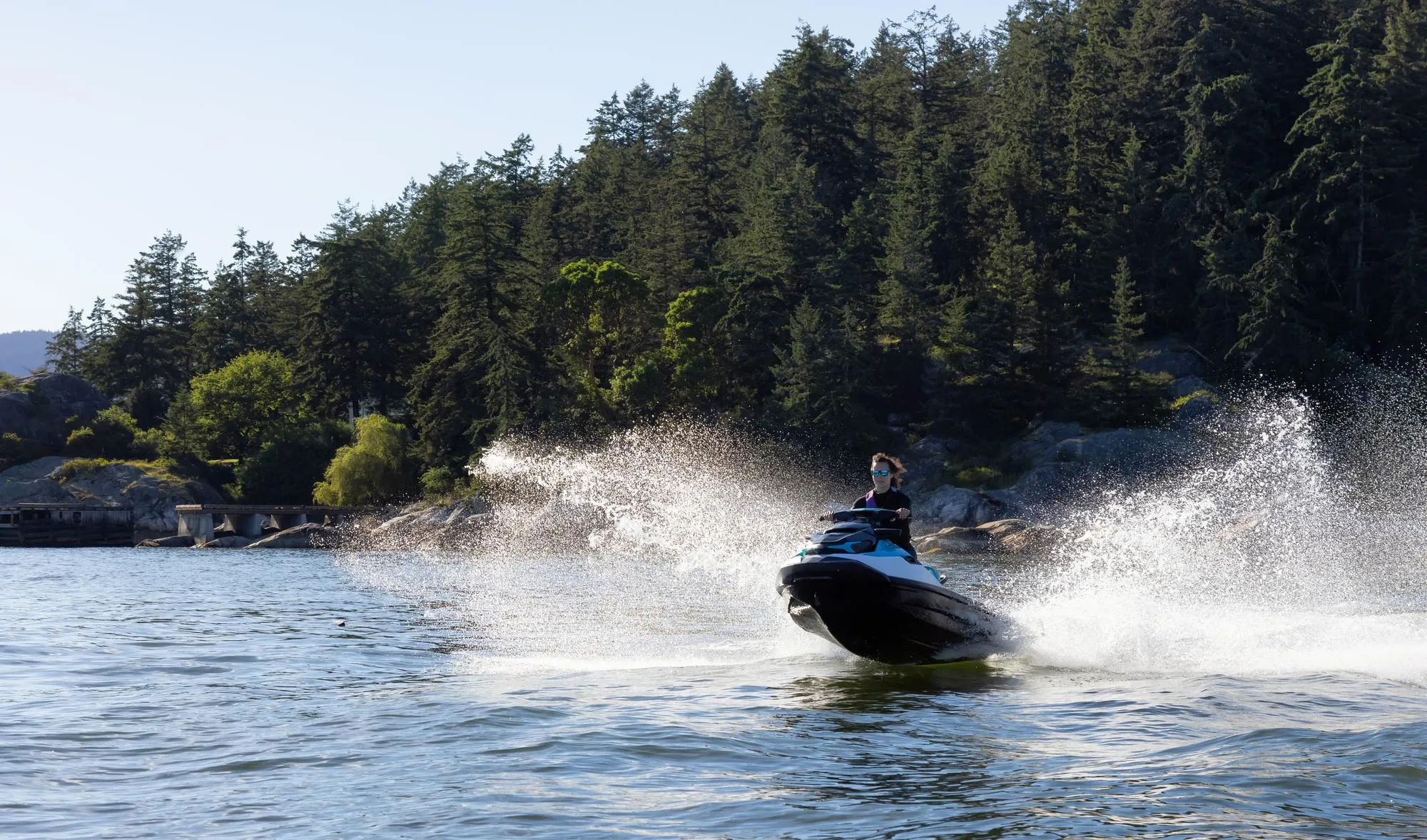 A person on a black and blue jet ski speeds across blue-green water, generating a large, sunlit white spray. A dense forest of evergreen trees lines the rocky shore in the background, with distant hills and wooden structures visible under a clear sky.