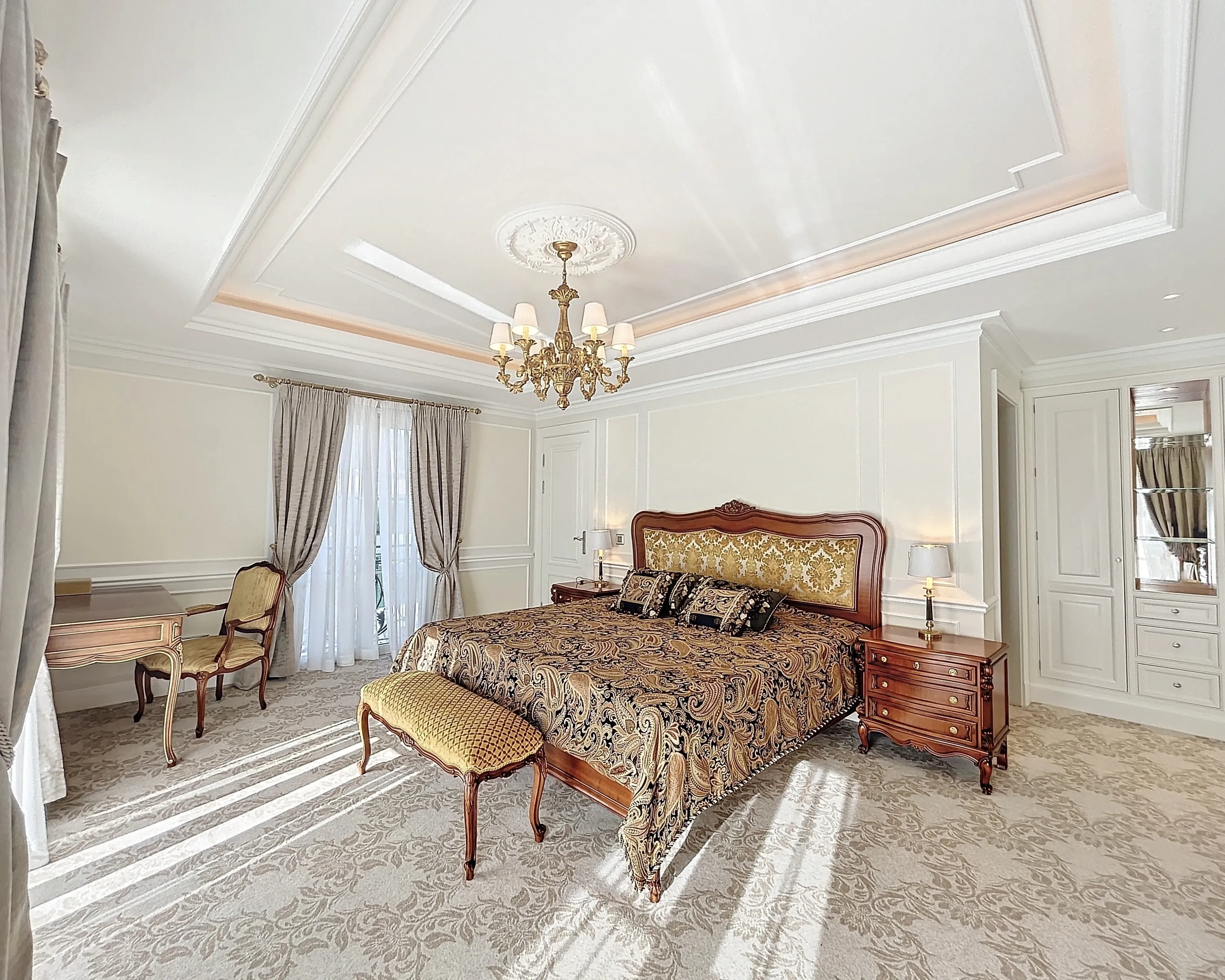 A luxurious bedroom showcases a large bed with a dark wood frame and a gold patterned headboard, covered by a black and gold paisley bedspread. Two dark wood nightstands with lamps flank the bed, and a gold upholstered bench sits at its foot. On the left, a matching dark wood desk and an ornate gold chair are positioned near grey and sheer white curtains. The room has light cream walls with panel molding, a multi-tiered ceiling with a central gold chandelier, and a light beige patterned carpet. Built-in white cabinets with mirrored sections line the right wall. Bright sunlight casts strong diagonal shadows across the carpet.