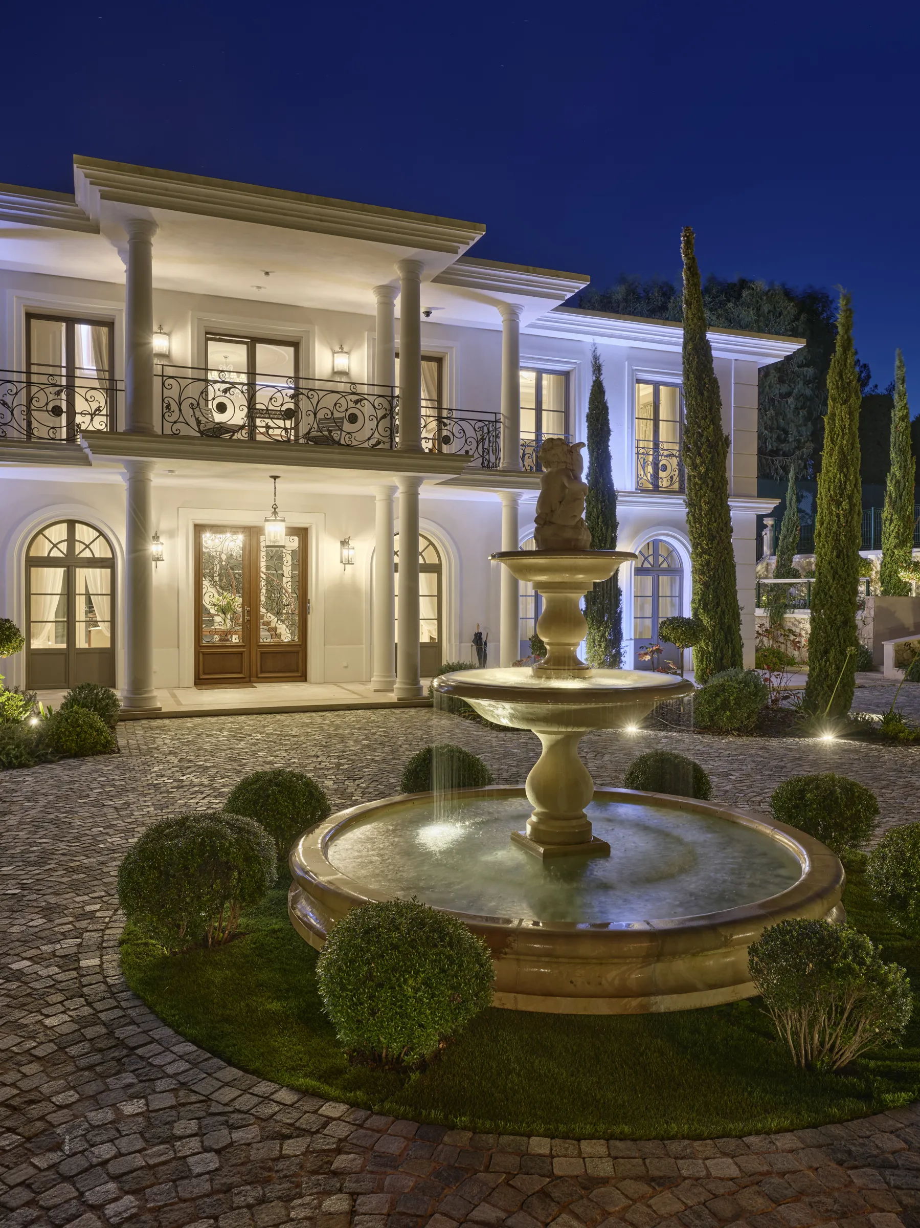An opulent, illuminated mansion with white columns and balconies featuring elaborate black ironwork stands under a deep blue, star-dotted night sky. In the foreground, a three-tiered stone fountain with a cherub figure flows, encircled by trimmed green bushes and a curved cobblestone driveway. Warm and cool lights highlight the architecture and landscaping.