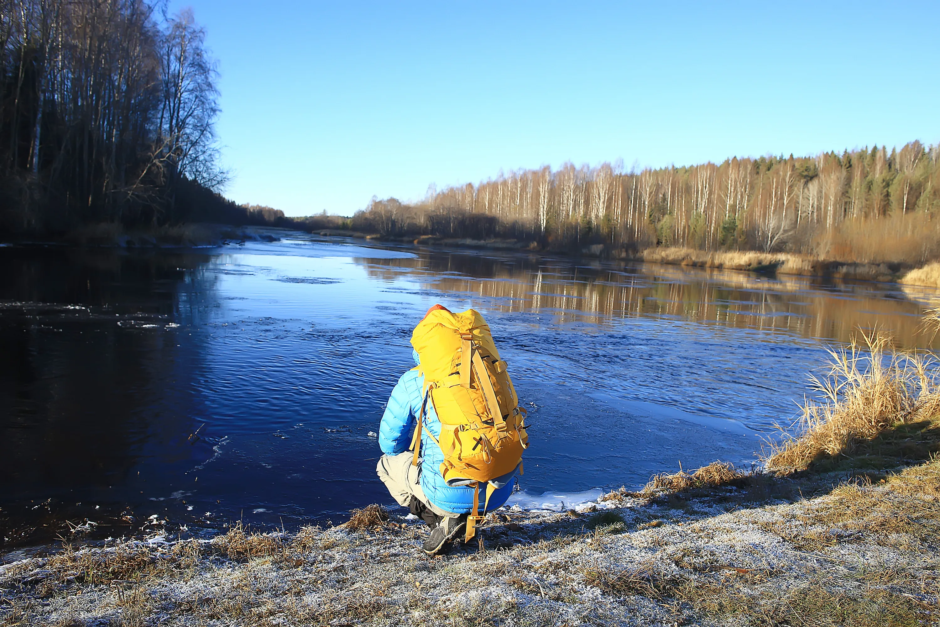 A person in a blue jacket and bright yellow backpack squats by a partially frozen river, observing the water under a clear blue sky. The foreground ground is covered in white frost, while the riverbanks are lined with bare trees and dry reeds, indicating a crisp, sunny late autumn or early winter day.