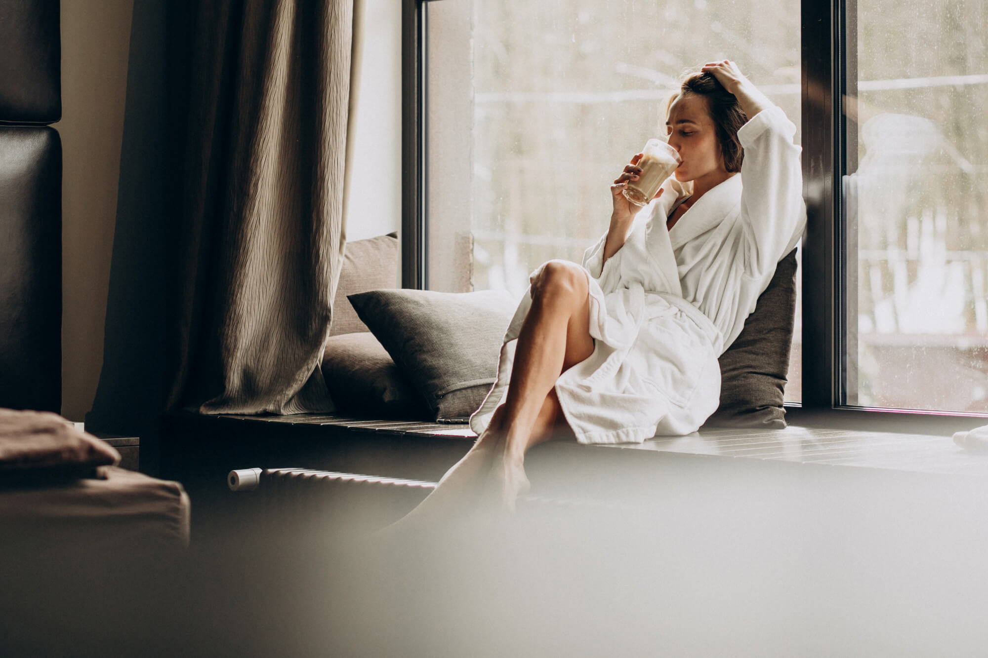 A woman in a white bathrobe sits on a window ledge, sipping a light brown drink from a glass. She is positioned by a large window, with grey pillows behind her and dark brown curtains to the left. Soft natural light enters through the window, revealing a blurred outdoor scene. A dark radiator is visible below the sill, and dark wall panels are on the far left.