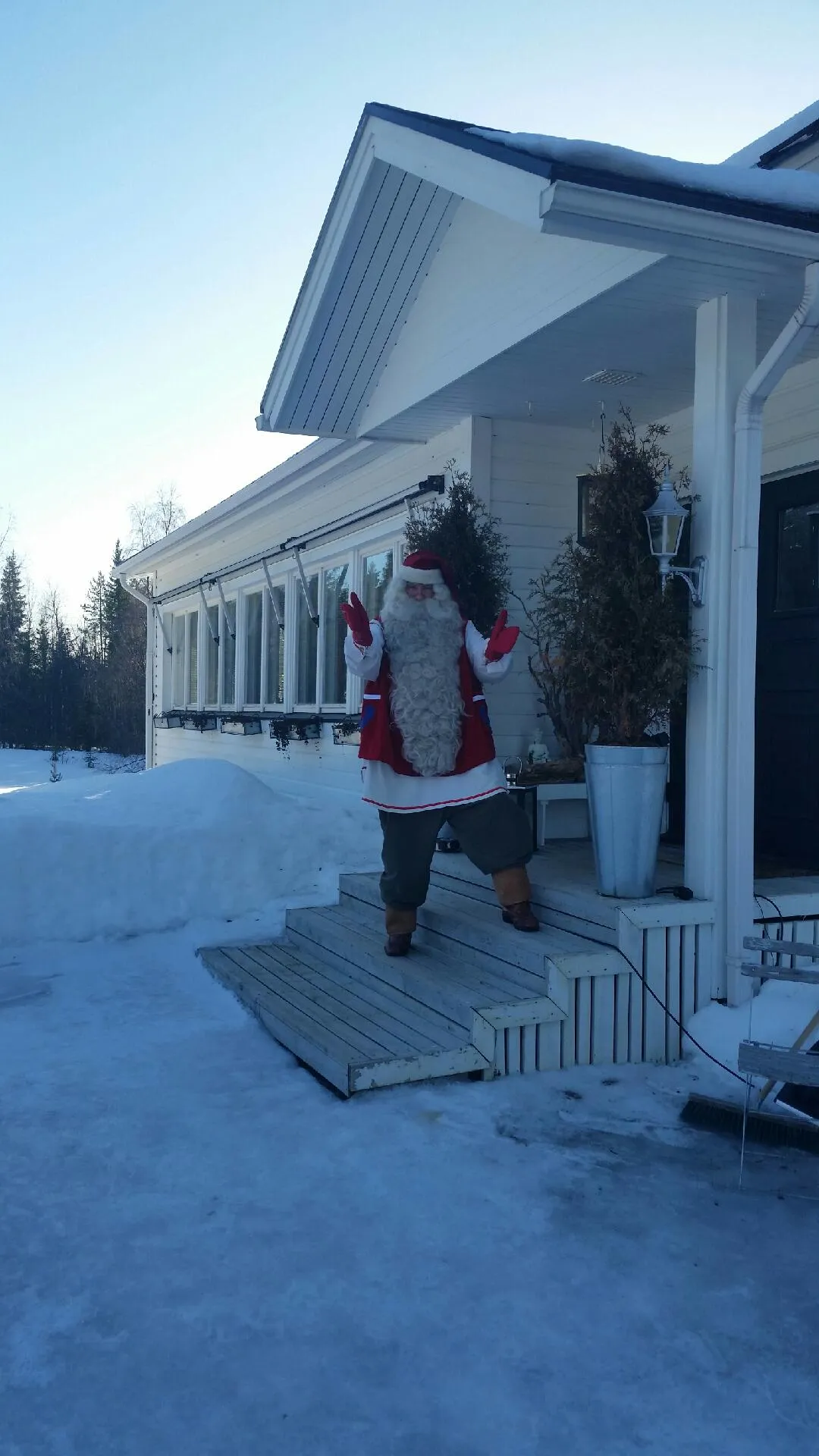 A person in a red and white Santa Claus costume with a long white beard stands on a wooden porch with steps, in front of a white house. The ground is covered in snow and ice, with evergreen trees visible in the background under a clear blue sky. The house has white siding, multiple windows, a dark roof with some snow, and a dark entrance door on the right.