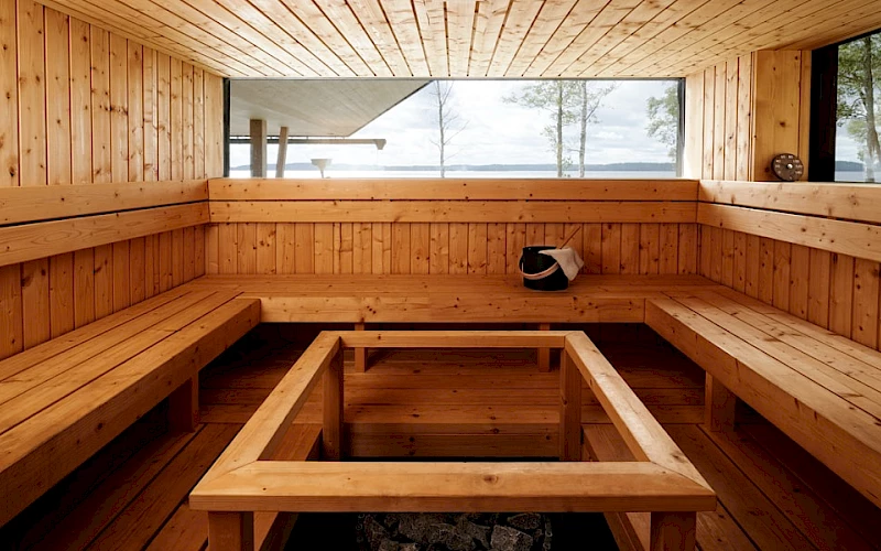 A bright, modern sauna interior crafted entirely from light-toned wooden planks. Tiered benches line the walls, and a large horizontal window offers a panoramic view of a lake, distant land, and trees under a cloudy sky. In the center, a square wooden frame surrounds a dark opening containing sauna rocks. A black bucket with a white cloth and ladle rests on a bench, and a round gauge is mounted on the right wall. Natural light illuminates the warm wooden surfaces.