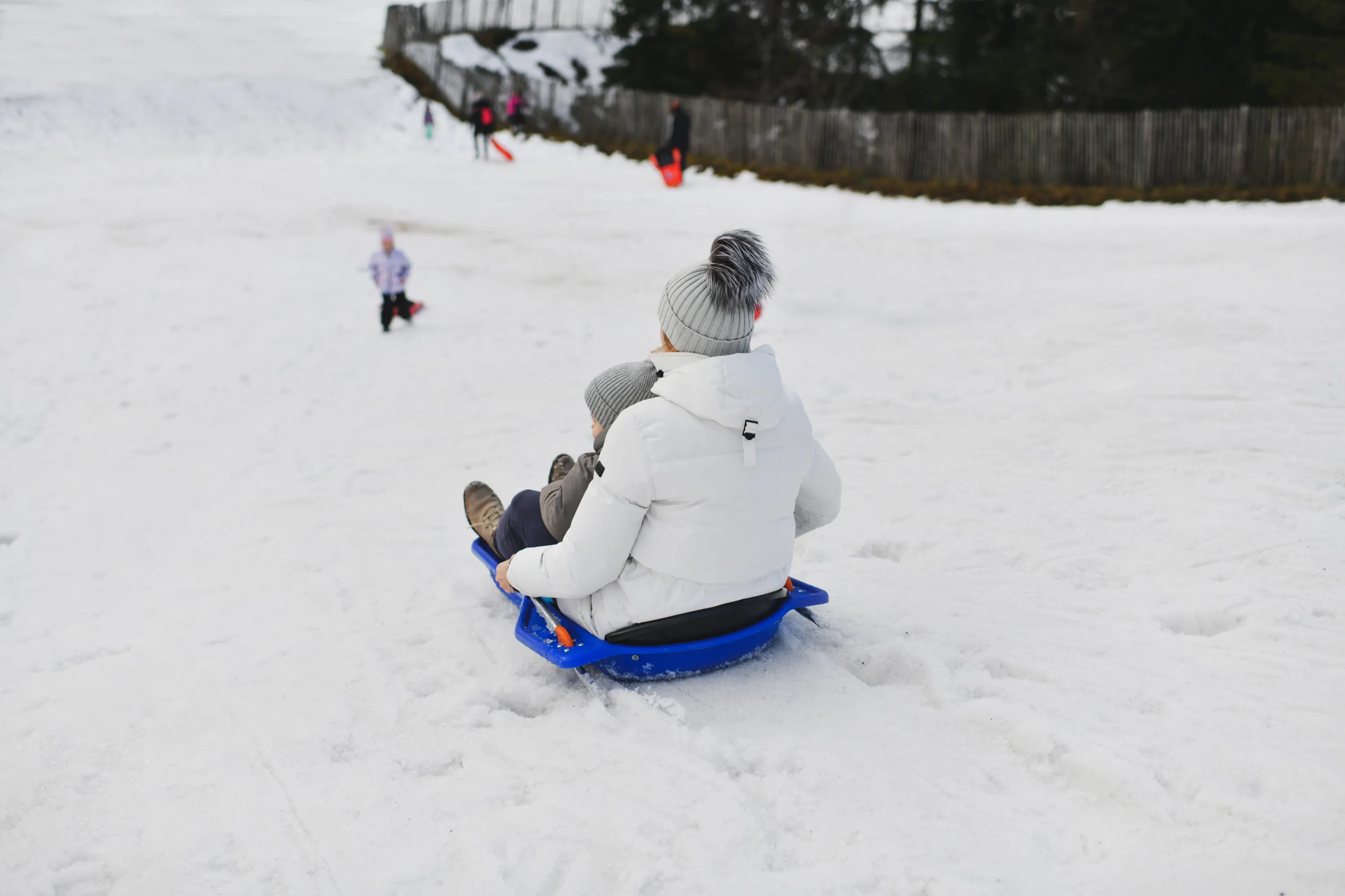 A person in a white puffer jacket and a grey pom-pom hat is sledding down a snow-covered hill with a child on a blue plastic sled. Both are seen from behind, with other blurry figures, a dark fence, and trees in the distant background. The winter scene is illuminated by soft, diffused daylight.