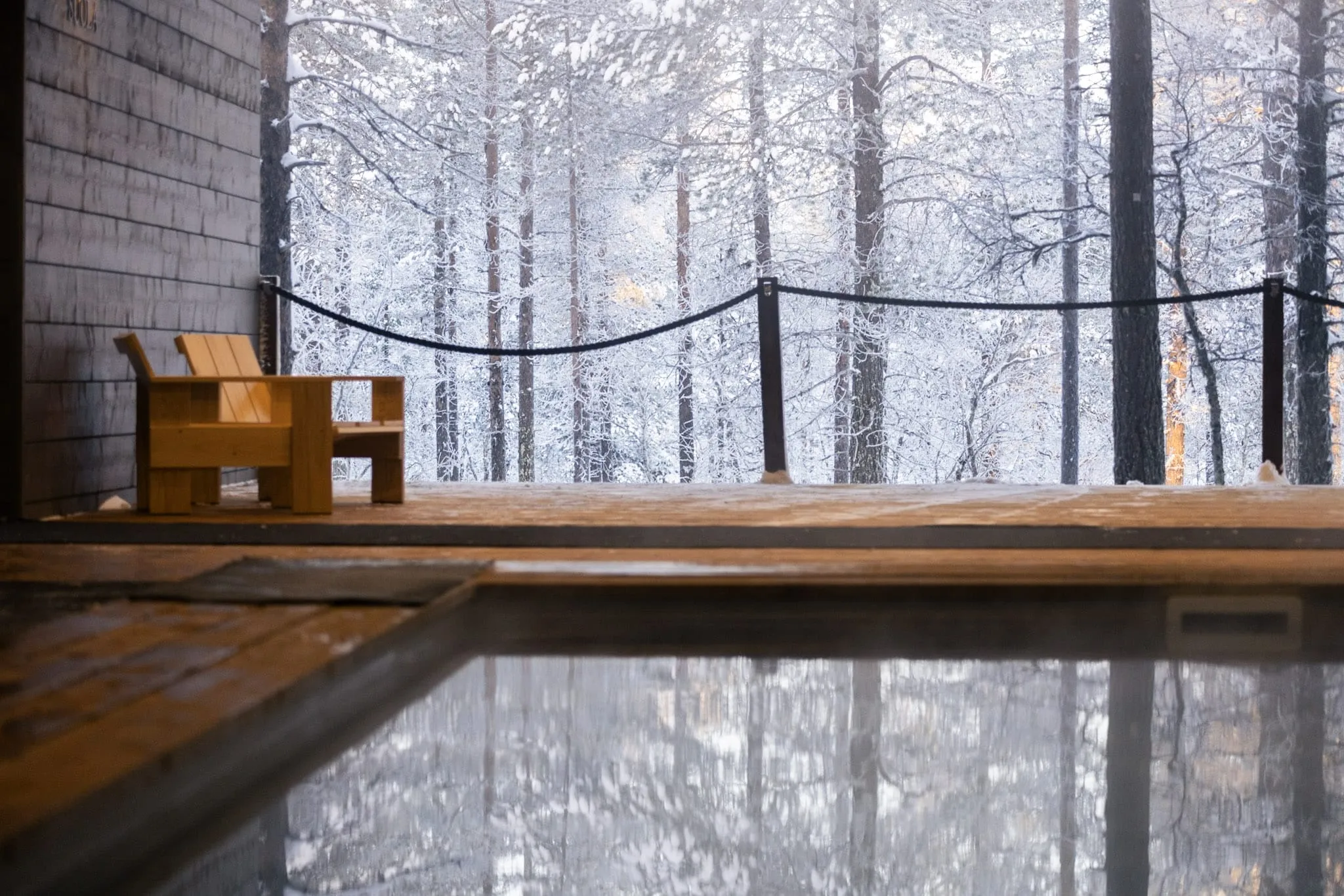 A dark gray wooden cabin wall stands beside a light brown wooden chair on a snow-dusted deck. A dark railing separates the deck from a dense, snow-covered forest of trees. In the foreground, a reflective pool mirrors the winter landscape under soft, diffused light.