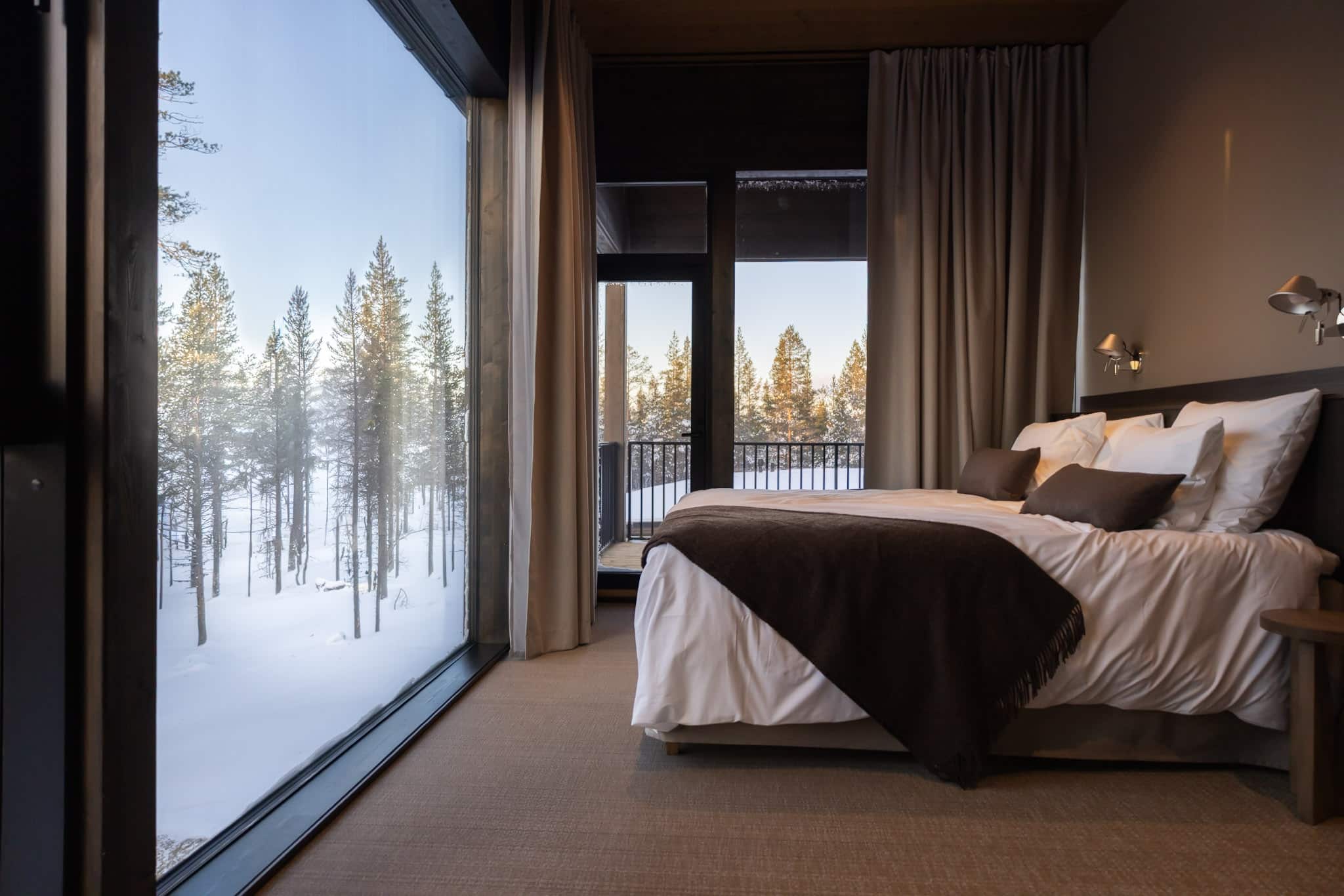 A modern bedroom features a large bed with white bedding, dark brown pillows, and a matching throw. A vast window on the left reveals a snowy pine forest under a clear blue sky, with sunlight filtering through the trees. In the center, a glass door opens to a balcony with a black railing, offering more views of the winter landscape. The room has light brown walls, a textured beige carpet, dark brown curtains, and two silver wall lamps above the bed.