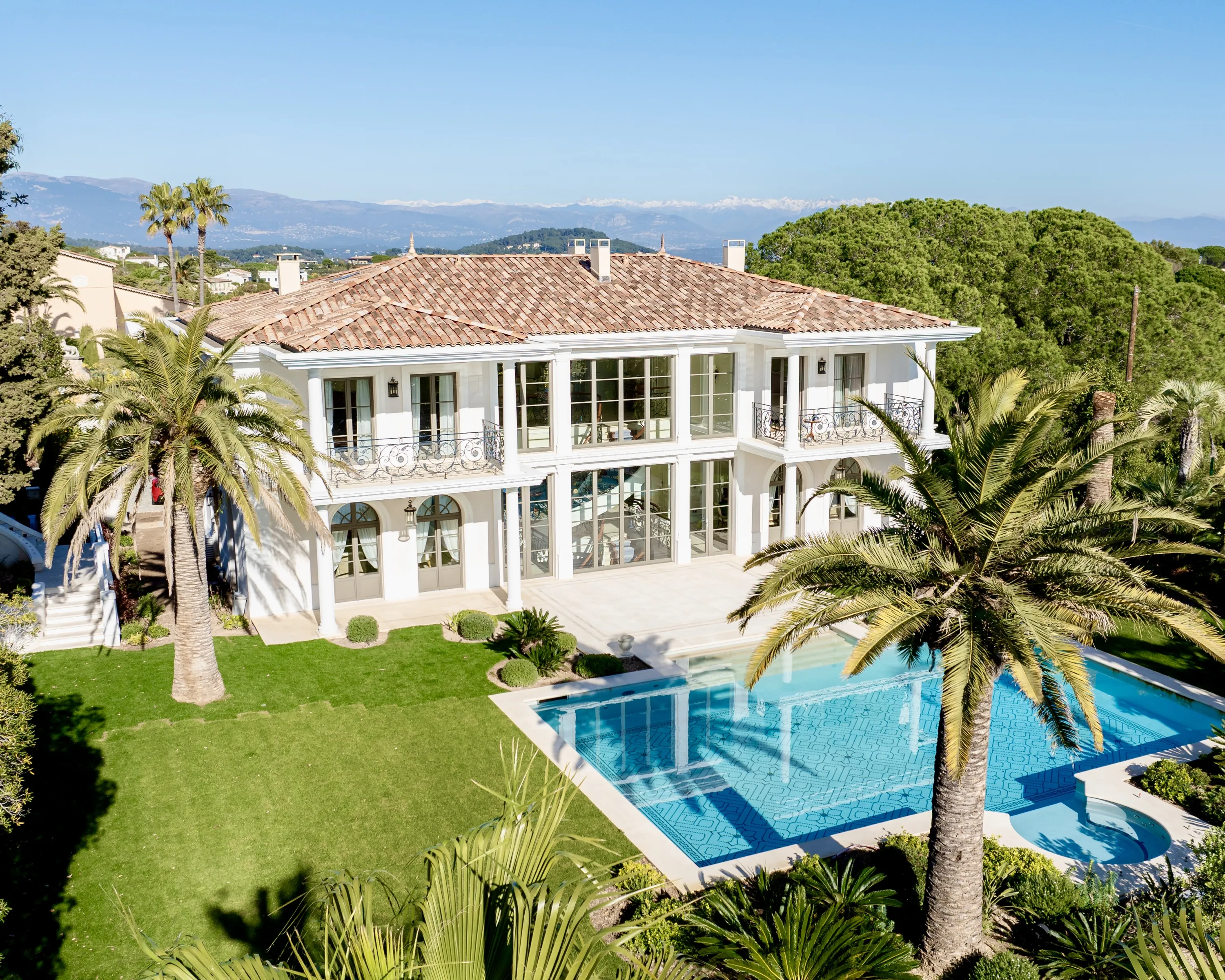 An aerial shot showcases a luxurious white villa with a terracotta roof, featuring large windows and balconies with elaborate black metal railings. In the foreground, a bright blue swimming pool with complex geometric blue mosaic patterns is set within a manicured green lawn, flanked by tall palm trees and various shrubs. The scene is bathed in bright sunlight, casting distinct shadows, with a clear blue sky above and distant snow-capped mountains on the horizon.