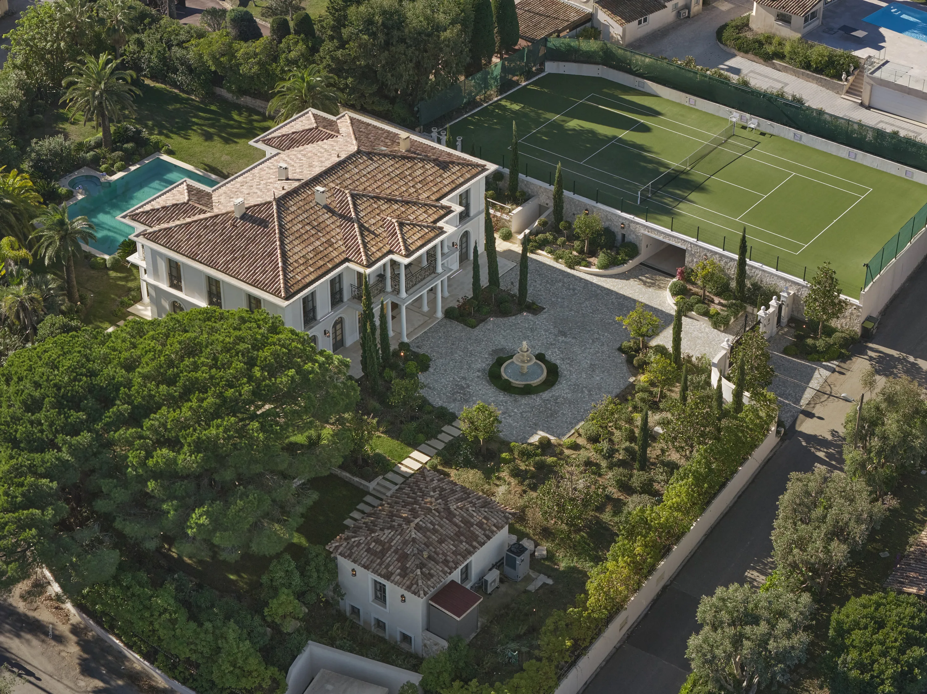 An aerial view captures a grand white villa with a terracotta tiled roof, featuring a blue swimming pool, a green tennis court, and a stone-paved circular driveway with a central fountain, all set within extensive green gardens under bright sunlight.