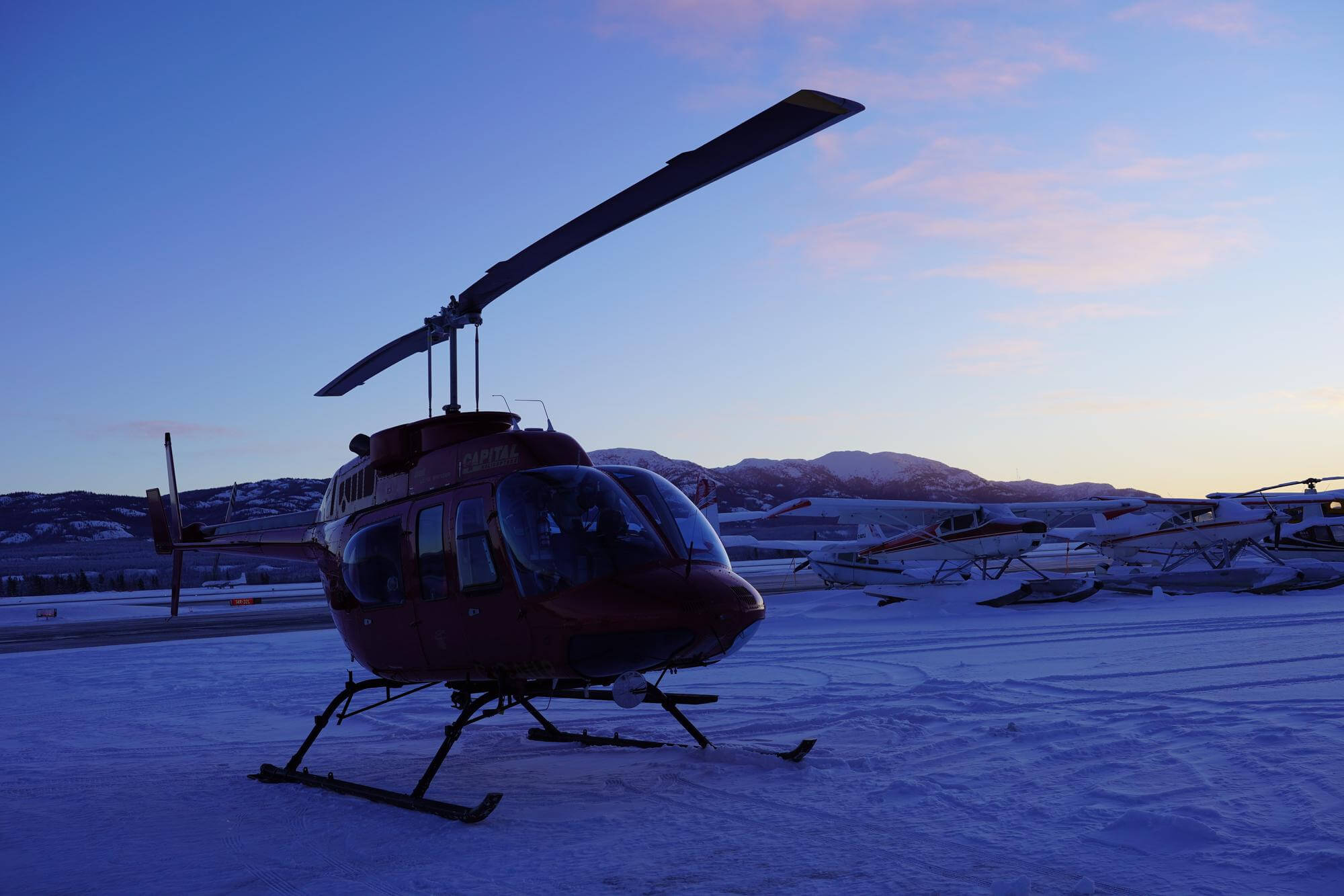 A red helicopter with "CAPITAL HELICOPTER" on its side rests on a snow-covered airfield. Its rotor blade points towards a twilight sky, which shows a gradient from deep blue to soft pink and orange. In the mid-ground, several small aircraft, some on floats, are parked and partially covered in snow, with snow-capped mountains visible in the distance.