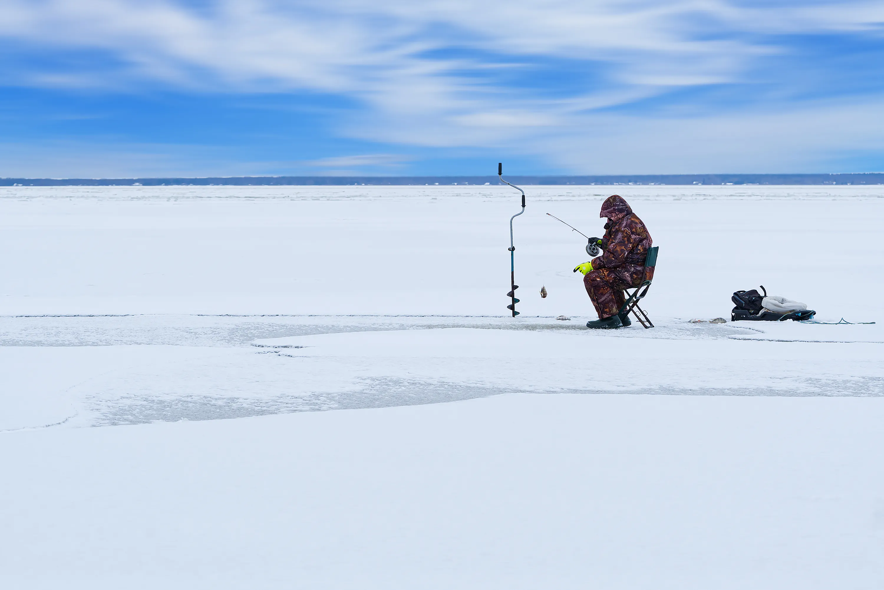 A person in a camouflage winter suit and yellow gloves sits on a green folding chair, ice fishing on a vast, snow-covered frozen lake. They hold a small fishing rod, with a fish caught on the line and another lying on the ice. An ice auger stands upright nearby, and a sled with gear is positioned to the right. A dark landmass stretches across the distant horizon under a bright blue sky with wispy white clouds.