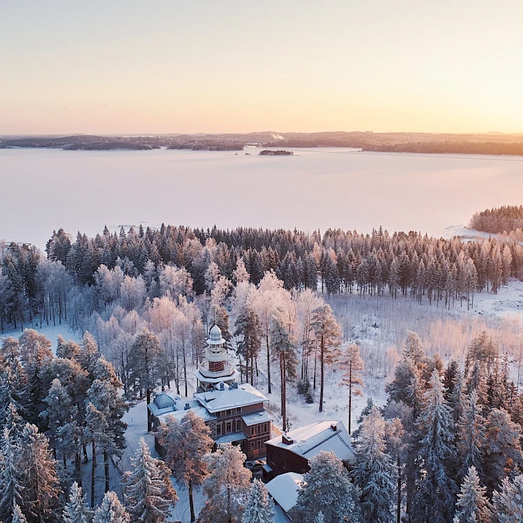 An aerial view captures a winter landscape at sunrise or sunset. A large building complex, featuring a prominent white-domed tower, is nestled within a dense forest of snow-covered trees. The trees, frosted white and pale pink, extend towards a vast, frozen lake reflecting the soft orange and pink hues of the sky. Distant land masses are visible across the icy water.