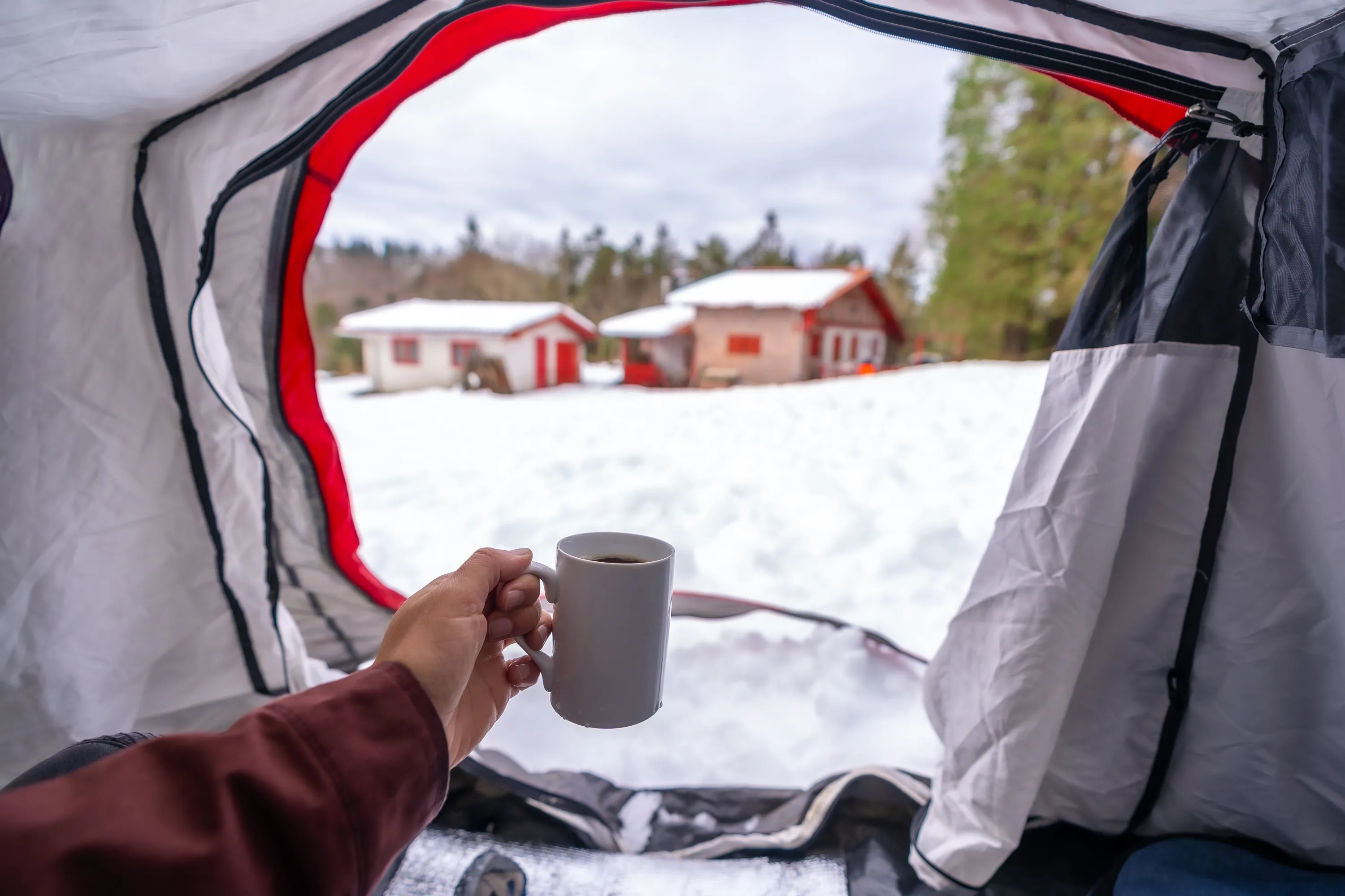 A first-person view from inside a white tent with black and red accents shows a hand holding a white mug of dark coffee. Beyond the tent opening, a snow-covered field leads to two small white houses with red roofs and doors, framed by distant trees under a cloudy sky. The lighting is soft and natural.