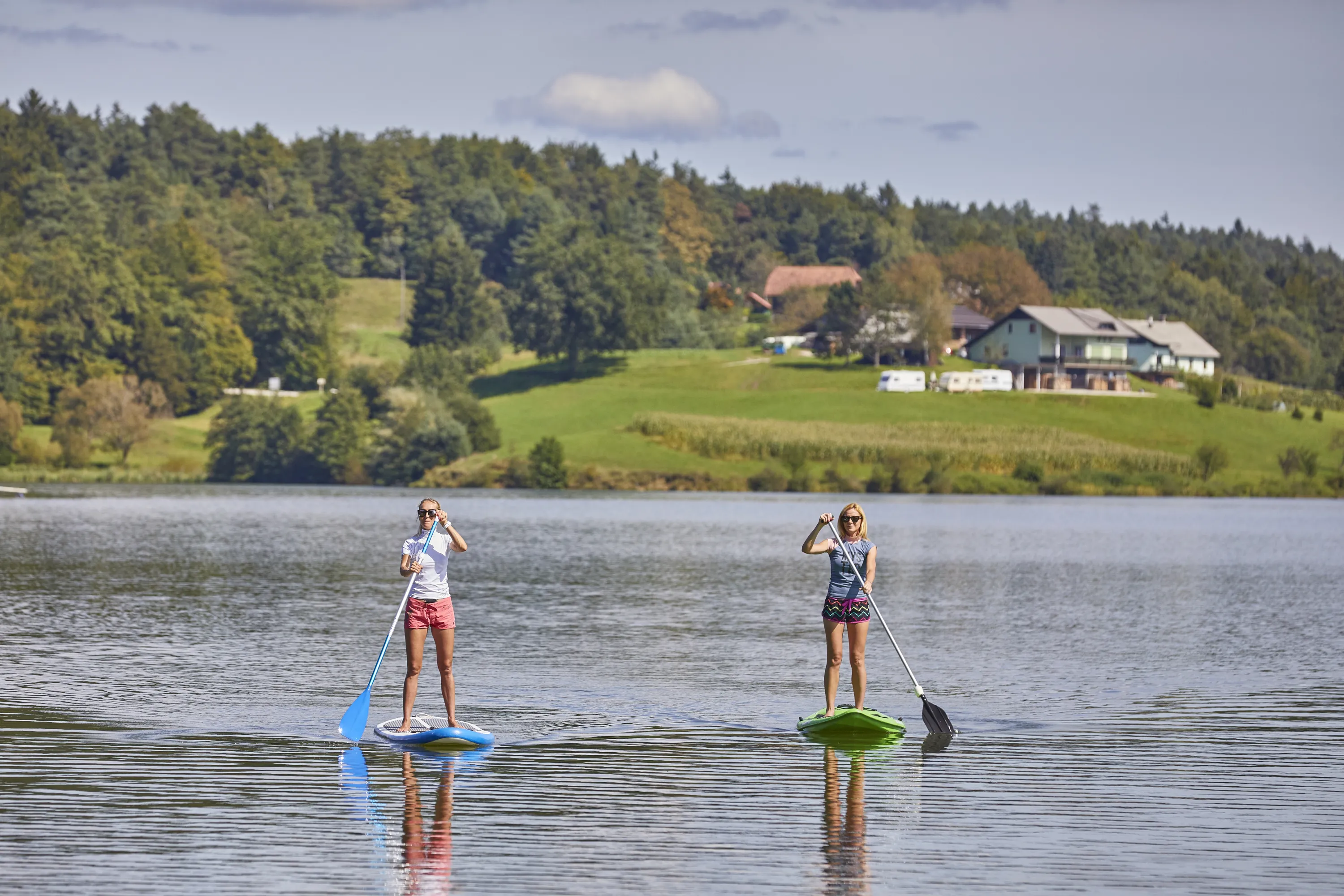 Two women are paddleboarding on a serene lake under a bright, partly cloudy sky. The woman on the left, wearing a white top and red shorts, stands on a blue and white paddleboard, holding a blue paddle. The woman on the right, in a grey top and patterned shorts, is on a green paddleboard, holding a silver and black paddle. Their reflections are visible on the calm water. In the background, lush green hills are covered with trees, and several houses with light-colored roofs are nestled among them, with a few white camper vans parked on a grassy area.